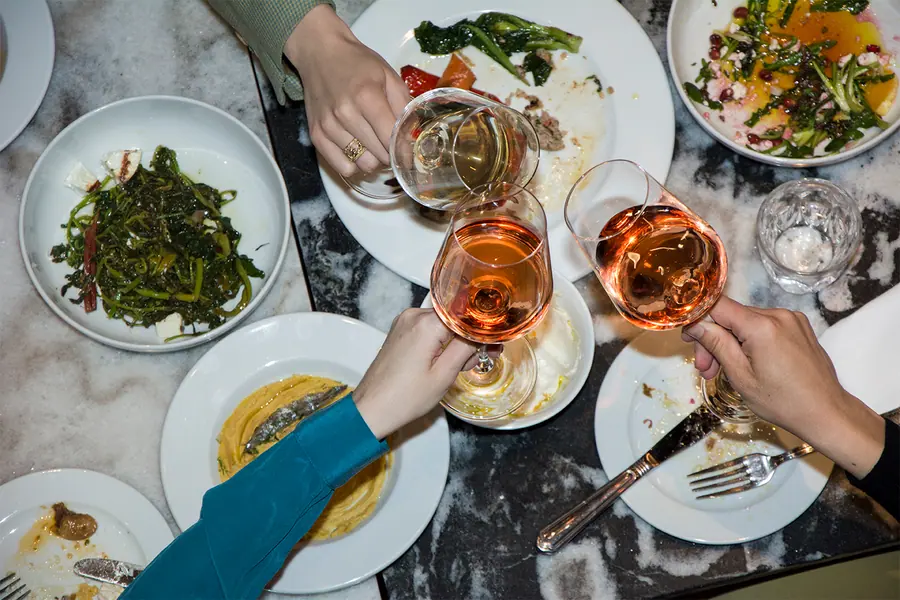 Hands clinking glasses of rosé over a table of shared plates